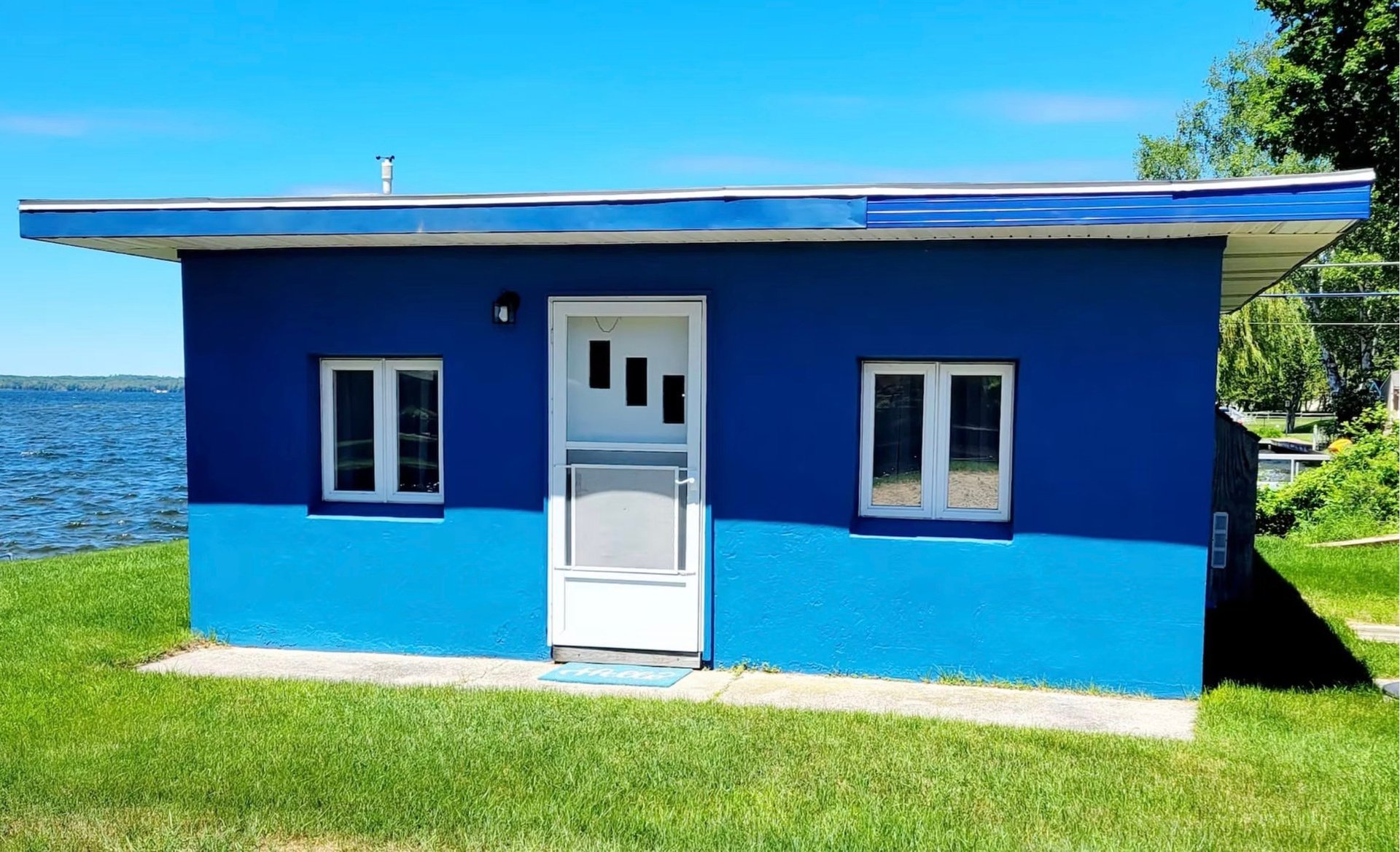Bright blue and white waterfront boathouse with white door and windows, situated on green grass near a lake