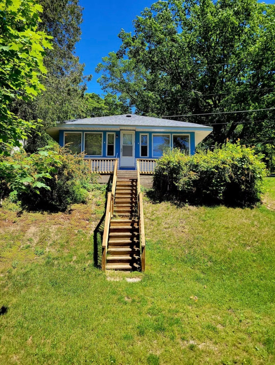 Blue cottage with white porch surrounded by green trees and landscaping, accessed by wooden stairs from a grassy yard