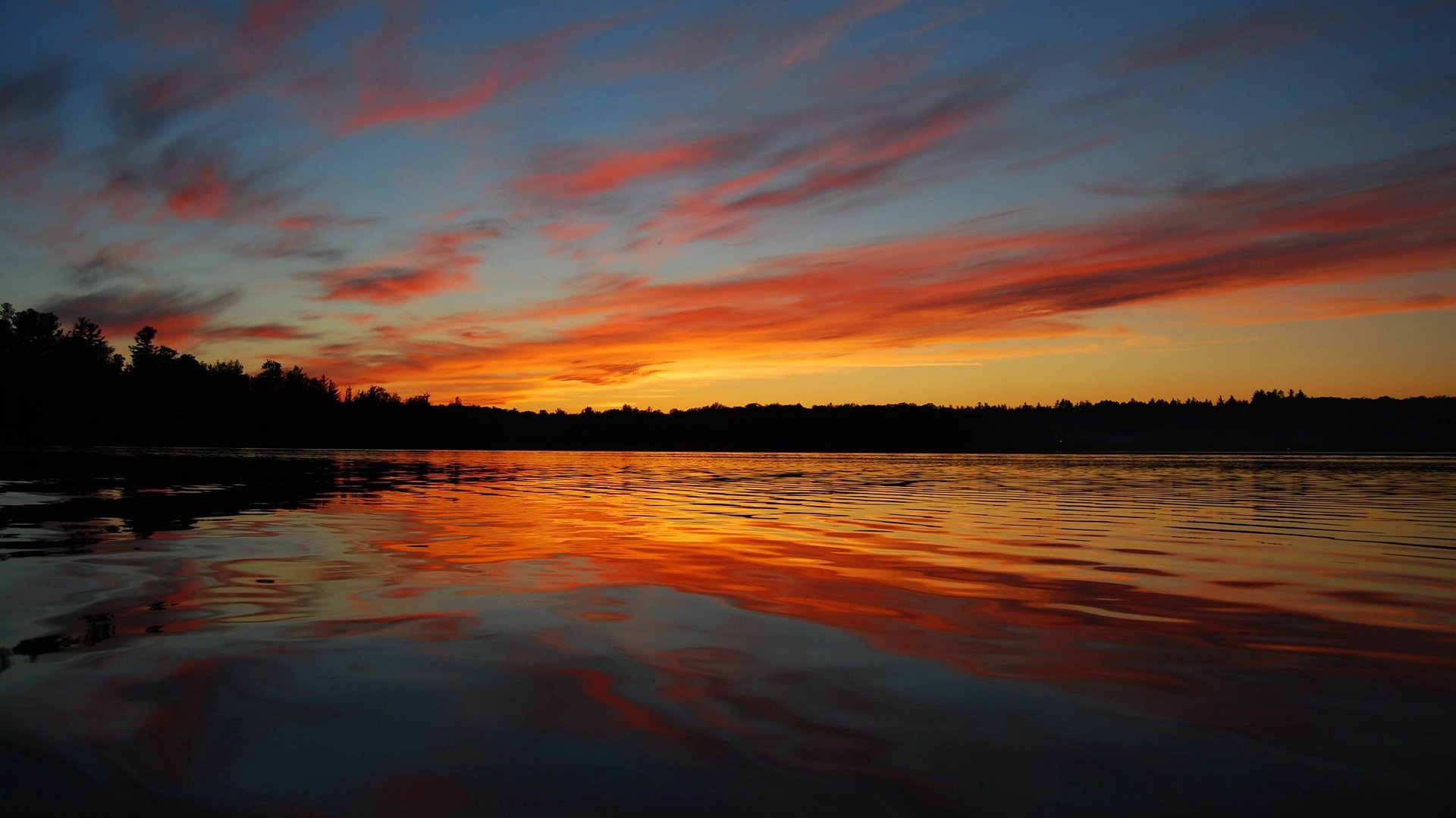 Sunset over calm water with orange and red sky reflecting on the lake surface, silhouetted trees along the shoreline