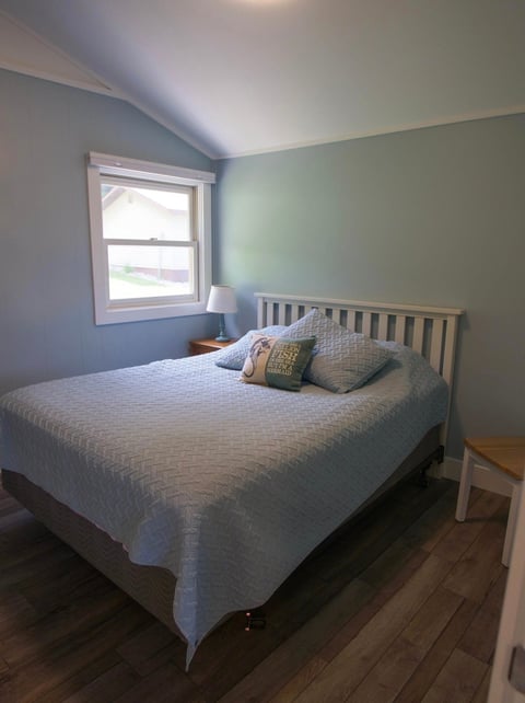 A bright bedroom with a white bed, blue-green walls, window with natural light, bedside table with lamp, and hardwood flooring