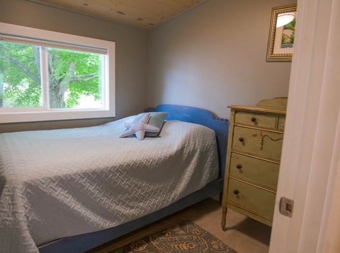 Cozy bedroom with blue-framed bed, gray bedding, wooden nightstand, and window with green outdoor view