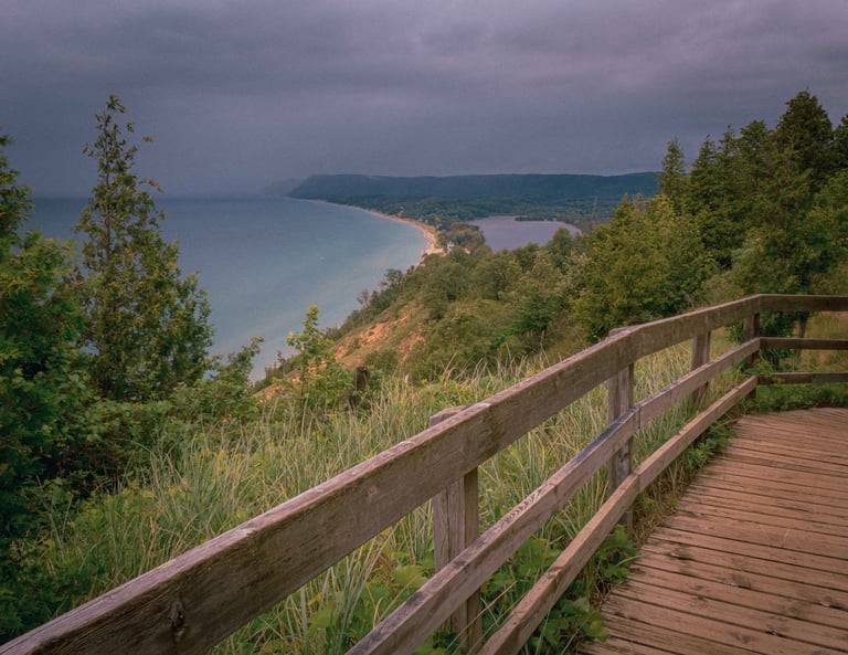 Wooden railing on a hiking trail overlook with panoramic view of a lake, sandy beach, forested shoreline, and distant mountains under a cloudy sky