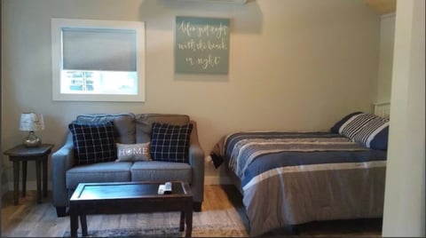 Studio apartment with blue couch, coffee table, bed with striped bedding, window, and Home sign decor