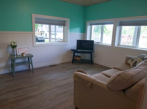 Bright living room with teal walls, tan sofa, TV stand, wooden floor, two windows, and beadboard wainscoting