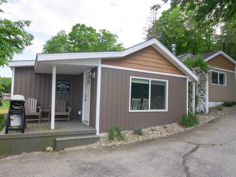 Brown and tan modular cabin with covered porch, white-trimmed windows, and white posts surrounded by trees