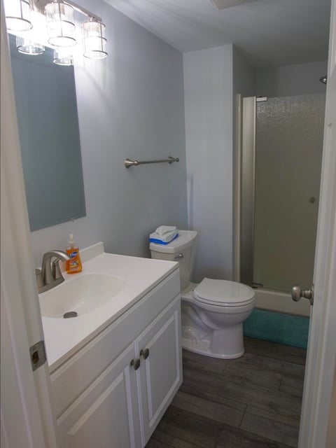 Modern bathroom with white vanity, toilet, and glass shower enclosure with blue-green tiled walls and dark wood flooring