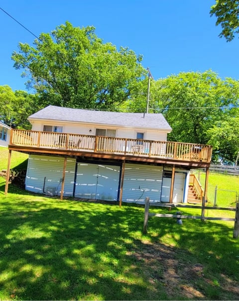 Two-story residential house with wooden deck and garage beneath, surrounded by green lawn and trees on a sunny day