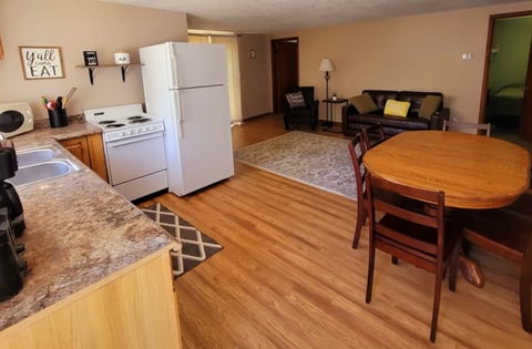 Open-concept apartment interior with kitchen area featuring white appliances and granite countertops, dining table, and living room with couch in background
