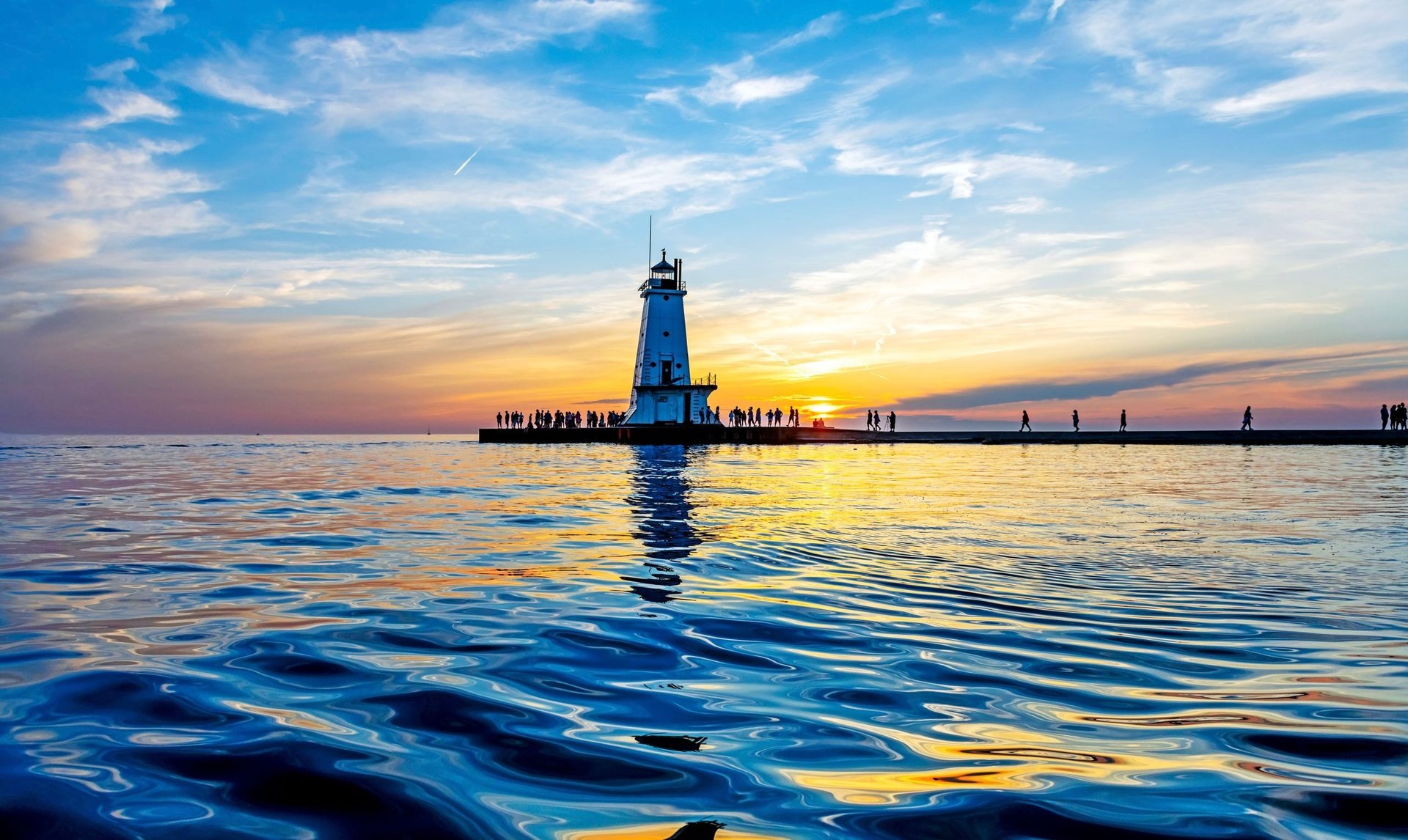 Blue lighthouse silhouetted against colorful sunset sky over calm water with sailboats in distance