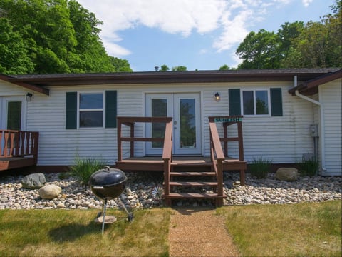 White mobile home with wooden deck and ramp, surrounded by gravel landscaping and green trees under blue sky