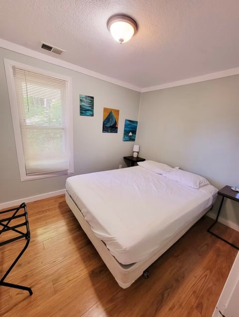 A minimalist bedroom with a white bed on a hardwood floor, window with blinds, small framed artwork on beige walls, and a ceiling light fixture