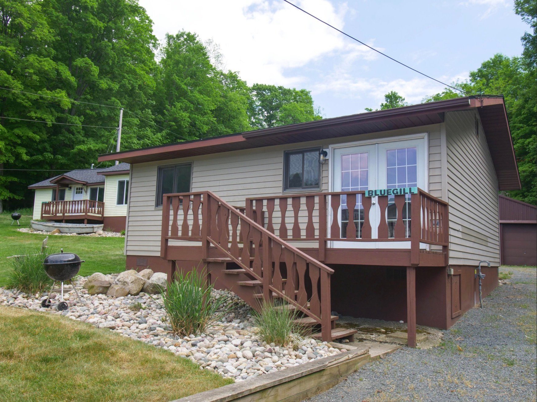 Brown and white mobile home with wooden deck and stairs, surrounded by green trees and landscaping with white gravel