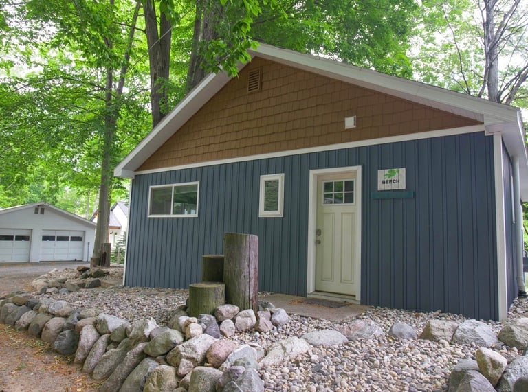 Modern small cabin with dark blue metal siding, tan roof, and cream door, surrounded by trees and gravel with stone foundation