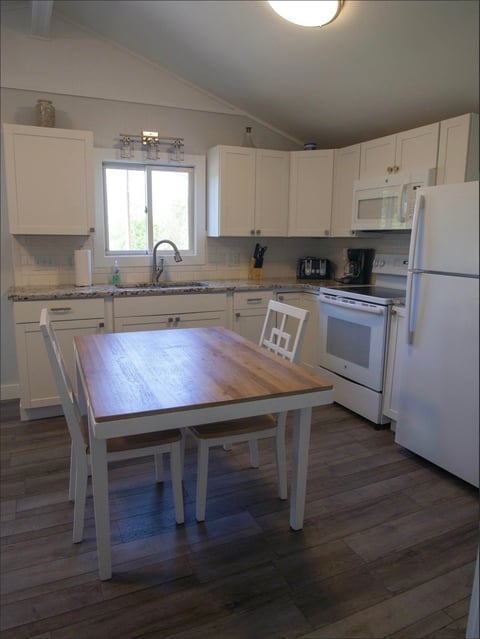 Bright kitchen with cream cabinets, white appliances, wood dining table with white chairs, and dark hardwood flooring