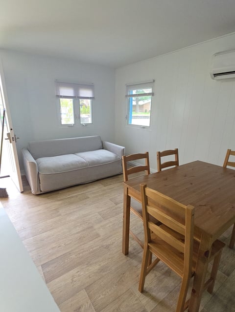 Bright apartment interior with wooden dining table and chairs, gray sofa, wood flooring, and windows with natural light