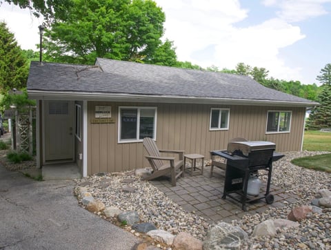 Beige single-story cabin with covered porch, Adirondack chairs, grill, and gravel patio surrounded by trees on a sunny day