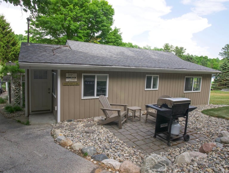Beige single-story cabin with covered porch, Adirondack chairs, grill, and gravel patio surrounded by trees on a sunny day