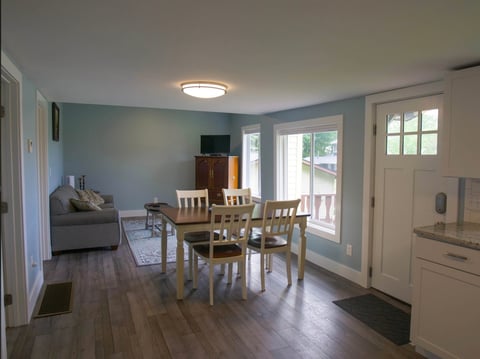 Interior of a light blue dining room with wooden table and chairs, hardwood floors, living area in background, and glass door entrance