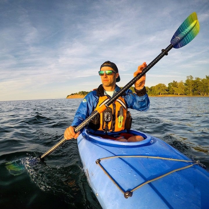 Man kayaking on blue kayak holding paddle, wearing yellow life vest and sunglasses on calm water with forested shoreline