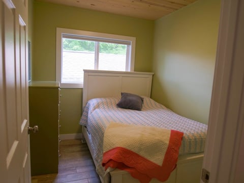 Small bedroom with yellow walls, single bed with white bedding, window with blinds, and wooden dresser