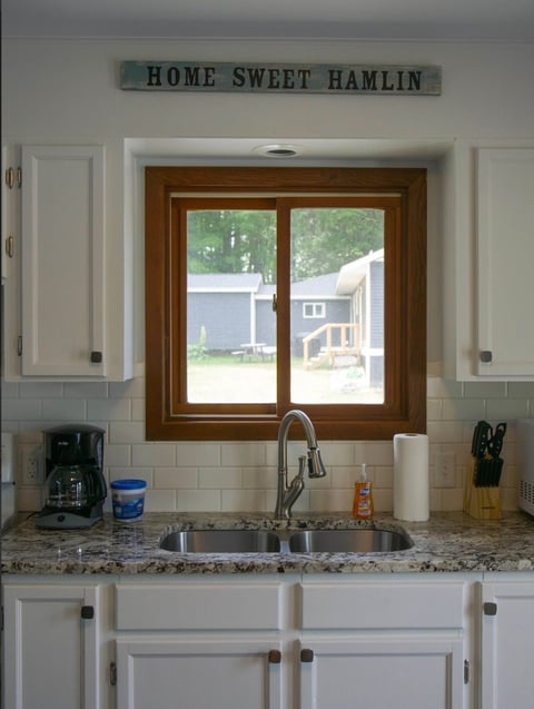 Kitchen window above sink with Home Sweet Hamlin sign, brown double-pane window, white cabinets, granite countertop, and chrome faucet