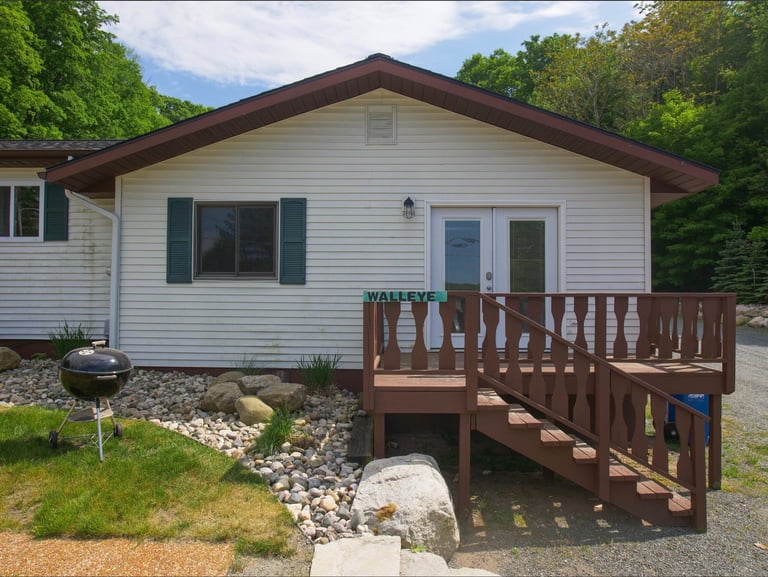 A beige mobile home with dark brown roof and wooden deck, surrounded by trees and landscaped with gravel and stones