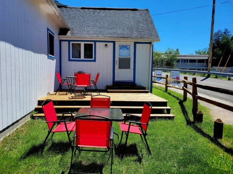 Small white and gray cabin with red chairs on grass, wooden deck, and rural ranch setting with wooden fencing