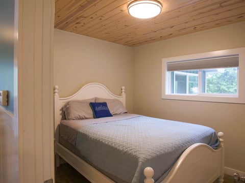 A bright bedroom with a white wooden bed, white bedding, and blue pillows. The room features wood ceiling, cream walls, and a window overlooking trees
