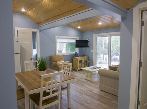 Modern open-concept living room with wood ceiling beams, light blue walls, wooden dining table, cream furniture, wall-mounted TV, and glass doors leading to patio
