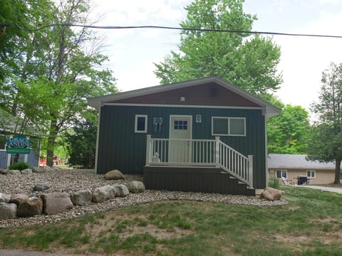 Dark green cabin with white railing porch surrounded by trees and gravel landscaping