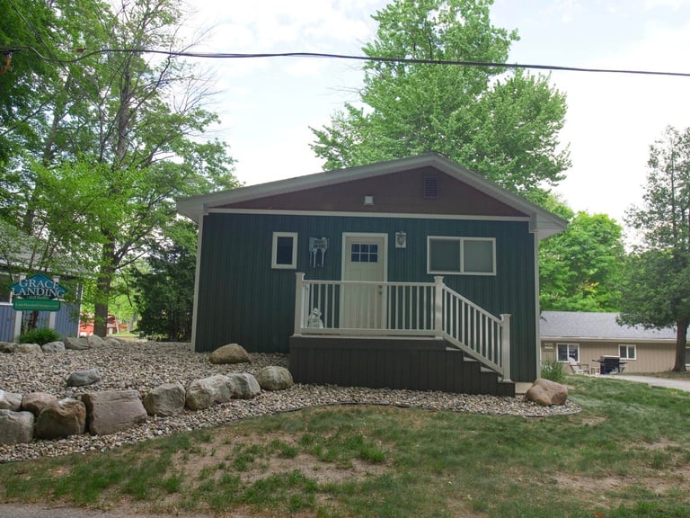 Dark green cabin with white railing porch surrounded by trees and gravel landscaping