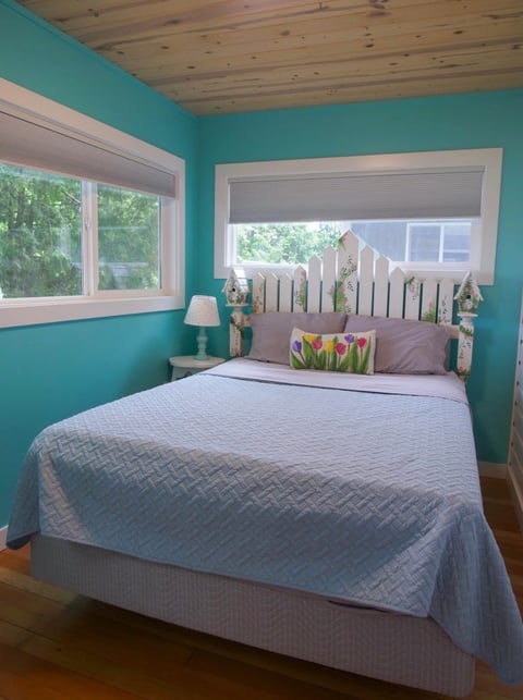 Bright turquoise bedroom with queen bed, white wooden headboard, and windows overlooking greenery