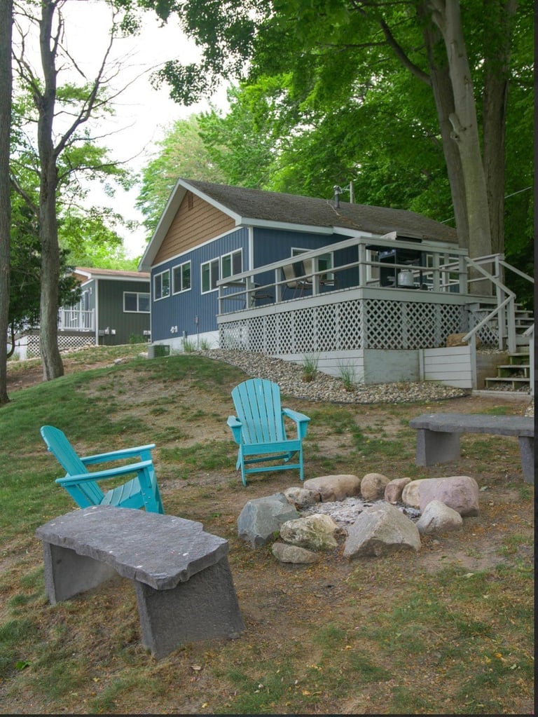 Blue cottage with deck surrounded by tall trees, featuring turquoise Adirondack chairs and fire pit in the yard