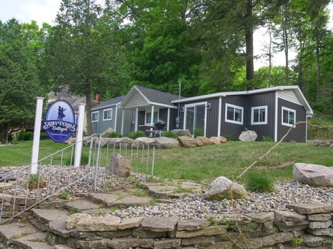 Dark blue and gray cottage with white trim beside a wooden sign, surrounded by trees and landscaped with rocks and stone retaining wall