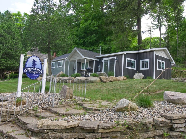 Dark blue and gray cottage with white trim beside a wooden sign, surrounded by trees and landscaped with rocks and stone retaining wall