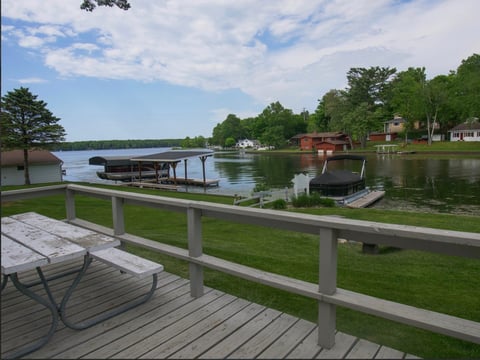 Wooden deck overlooking a calm river with boats, green grass, and trees on the shoreline under a partly cloudy sky