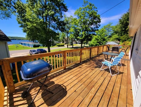 Wooden deck overlooking a lake with grill, chairs, and blue car parked below on a sunny day