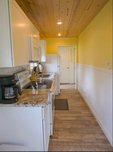 Bright yellow and white galley kitchen with white appliances, granite countertop, wooden ceiling, and wood flooring leading to hallway