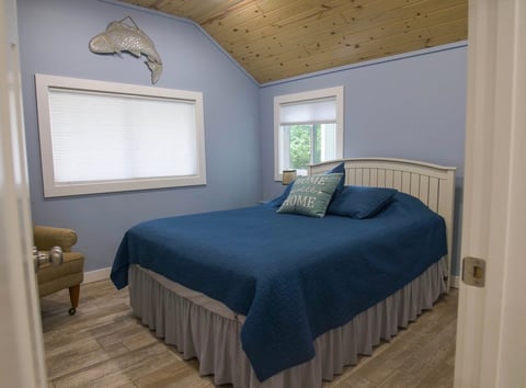 Cozy bedroom with blue walls, wooden vaulted ceiling, queen bed with dark blue bedding, two windows, and a fishing decal on the wall