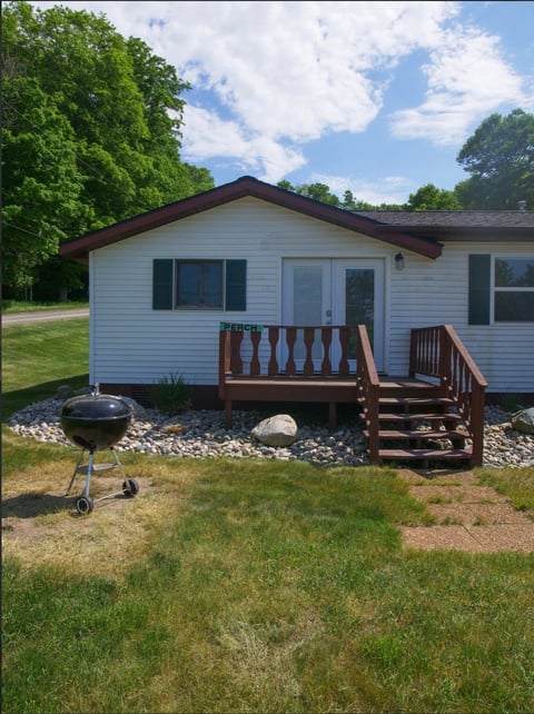 White single-wide mobile home with dark roof, wooden deck, and grill on a sunny day with green lawn and trees