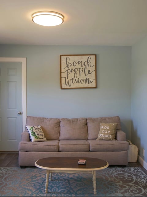 Cozy living room with a gray couch, wooden beach-themed wall sign, ceiling light, and decorative coffee table on a blue area rug