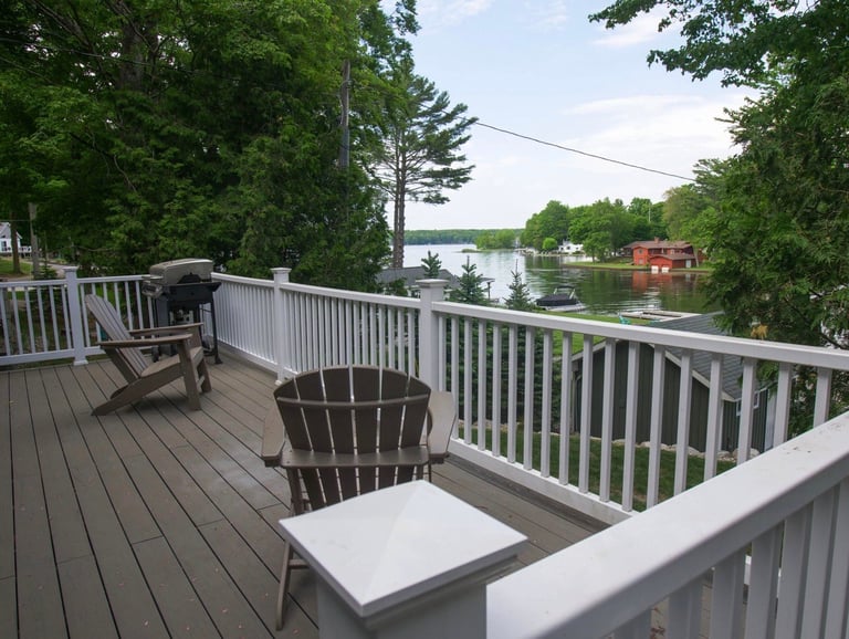 Waterfront deck with white railings, Adirondack chairs, and a view of a calm river lined with trees and houses