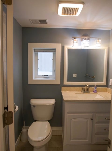 Modern bathroom interior with white toilet, vanity sink cabinet, framed mirror, window, and dark gray accent wall