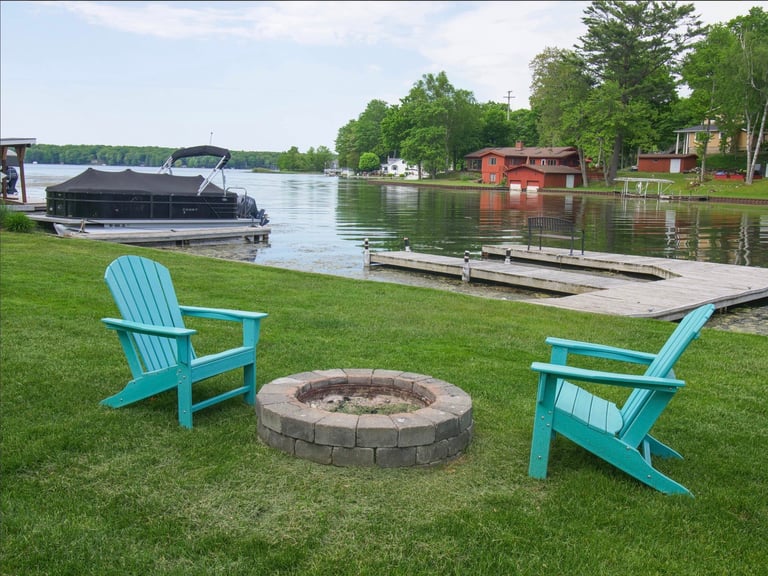 Two turquoise Adirondack chairs facing a fire pit on a lawn overlooking a waterfront dock and red boathouse