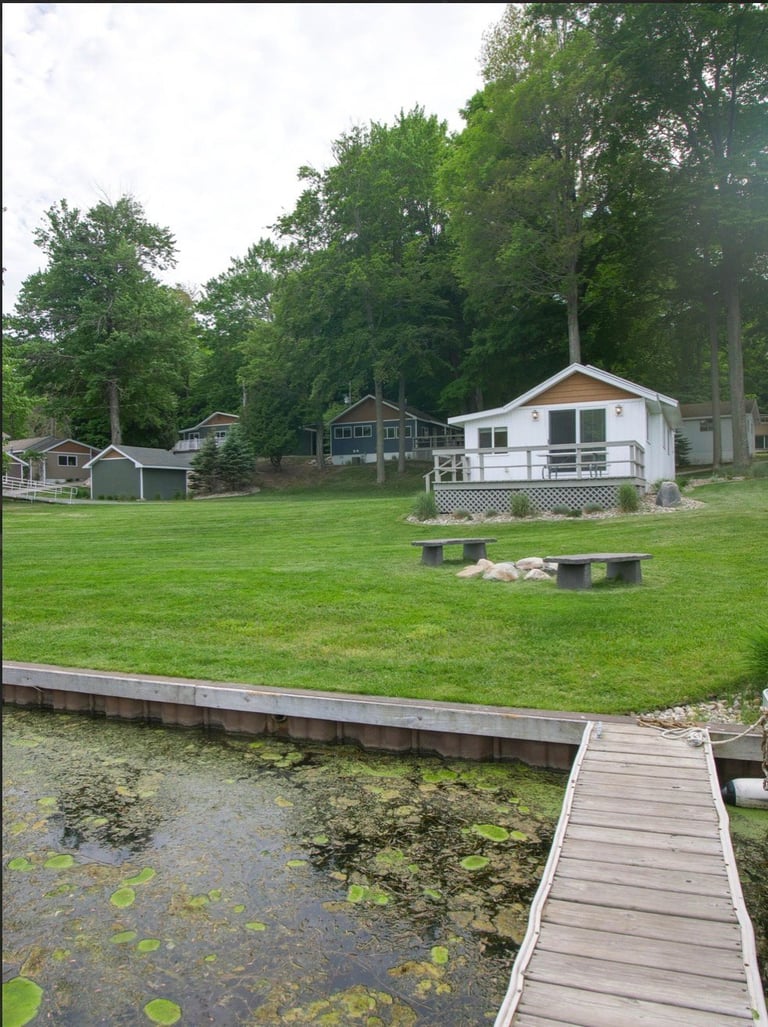 Waterfront cottage with white porch and deck on grassy lawn backed by tall trees, with wooden dock extending into small pond