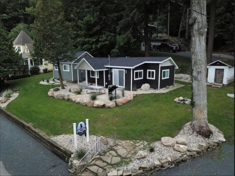 Dark gray cottage-style home on waterfront lot with manicured lawn, stone landscaping, and large tree in foreground