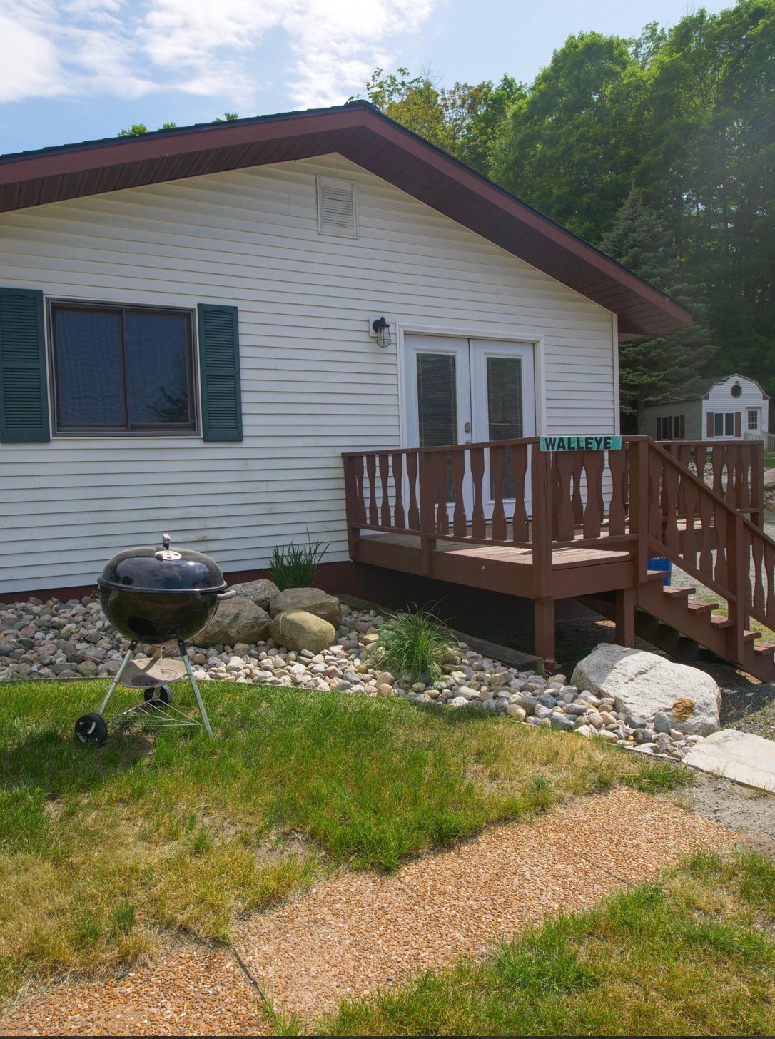 Single-story cottage with white siding, dark roof, brown deck, and gravel landscaping with a grill in front, surrounded by green trees