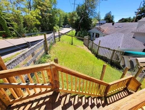 Wooden deck with stairs overlooking a green sloped yard, stone wall, and residential houses on a sunny day