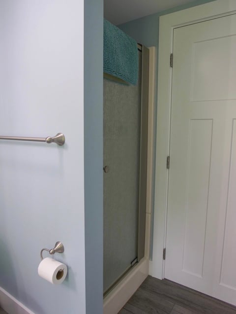 Bathroom interior with walk-in shower enclosure, white walls, toilet paper holder, towel bar, and white door visible on the right