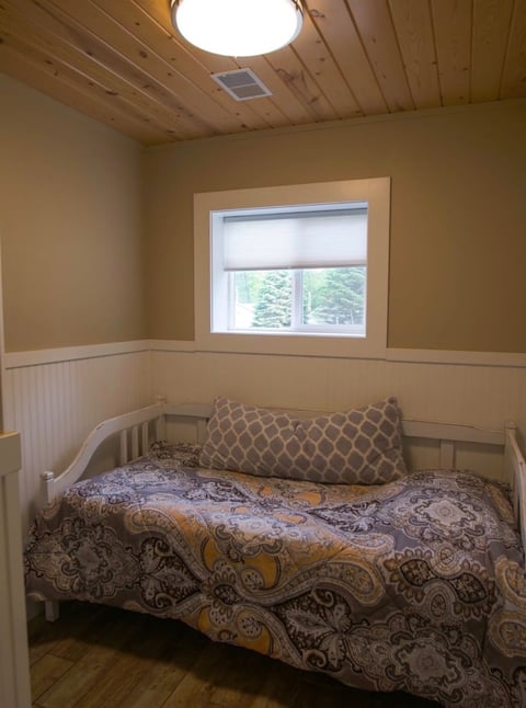 A simple bedroom with a white bed frame, patterned bedding, a tan accent pillow, wooden floor, and a large window with natural light